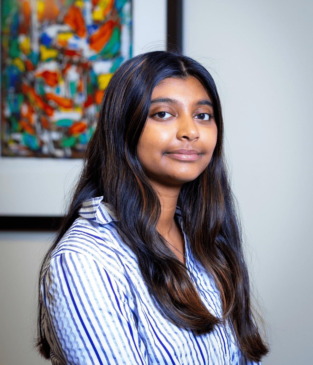 Portrait of a young woman with long dark hair wearing a white and blue striped shirt, seated indoors and looking at the camera with a slight smile, with a colorful abstract painting blurred in the background.