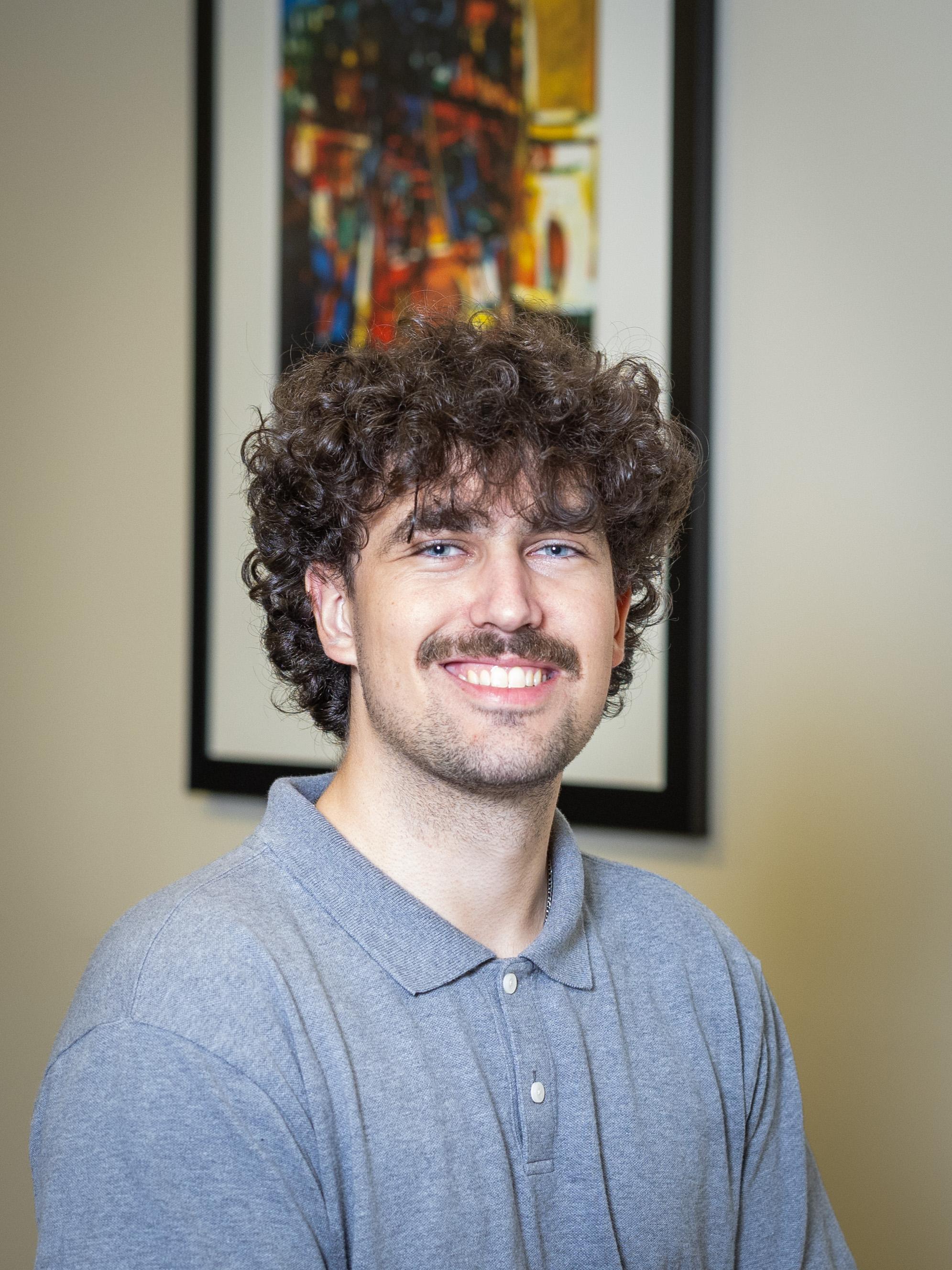 Portrait of a young man with curly brown hair and a mustache, smiling at the camera while wearing a light gray polo shirt, with a colorful abstract painting blurred in the background.  Do