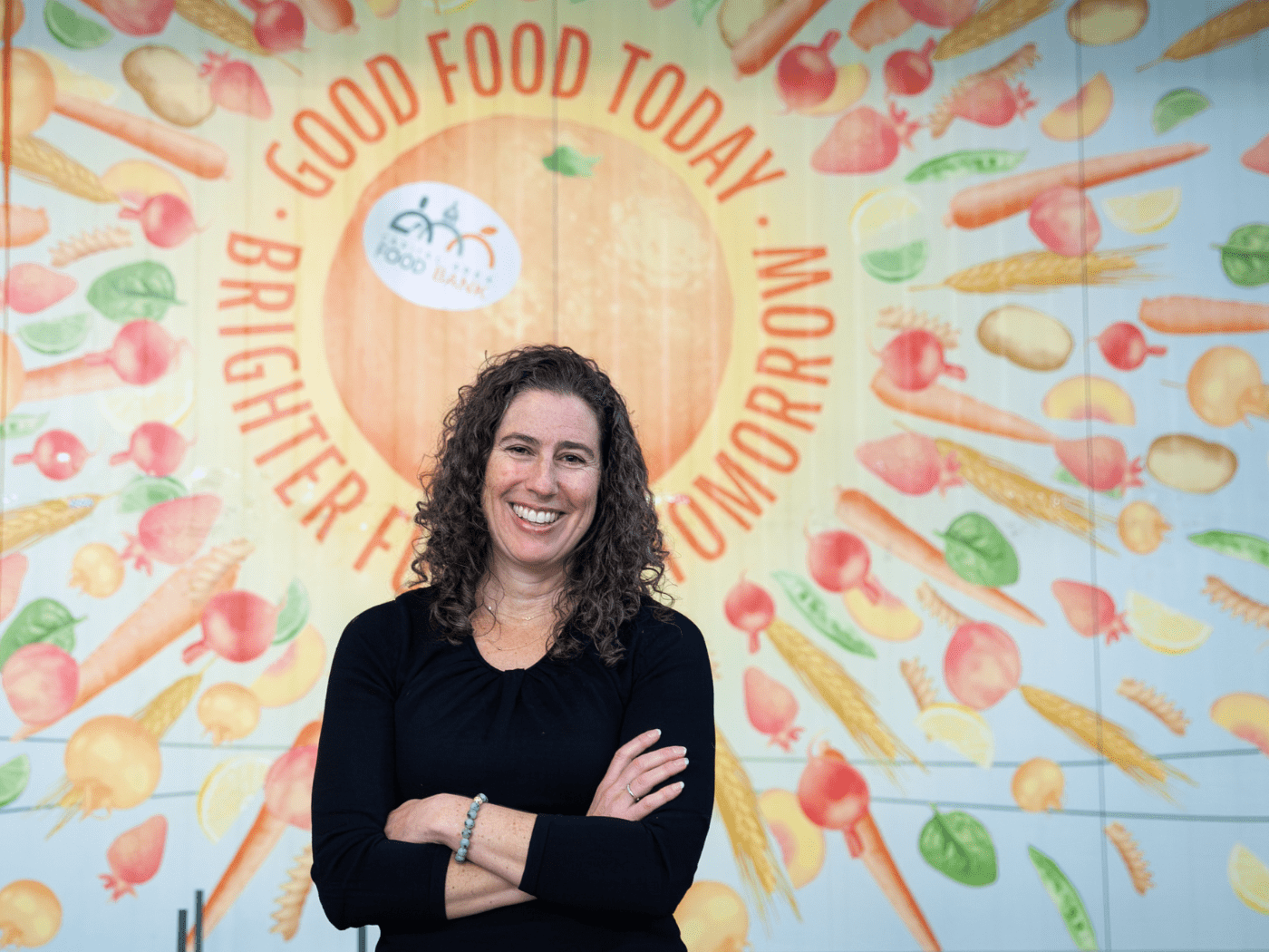White woman with curly long hair in a black top smiles in front of a wall mural with vegetables saying GOOD FOOD TODAY