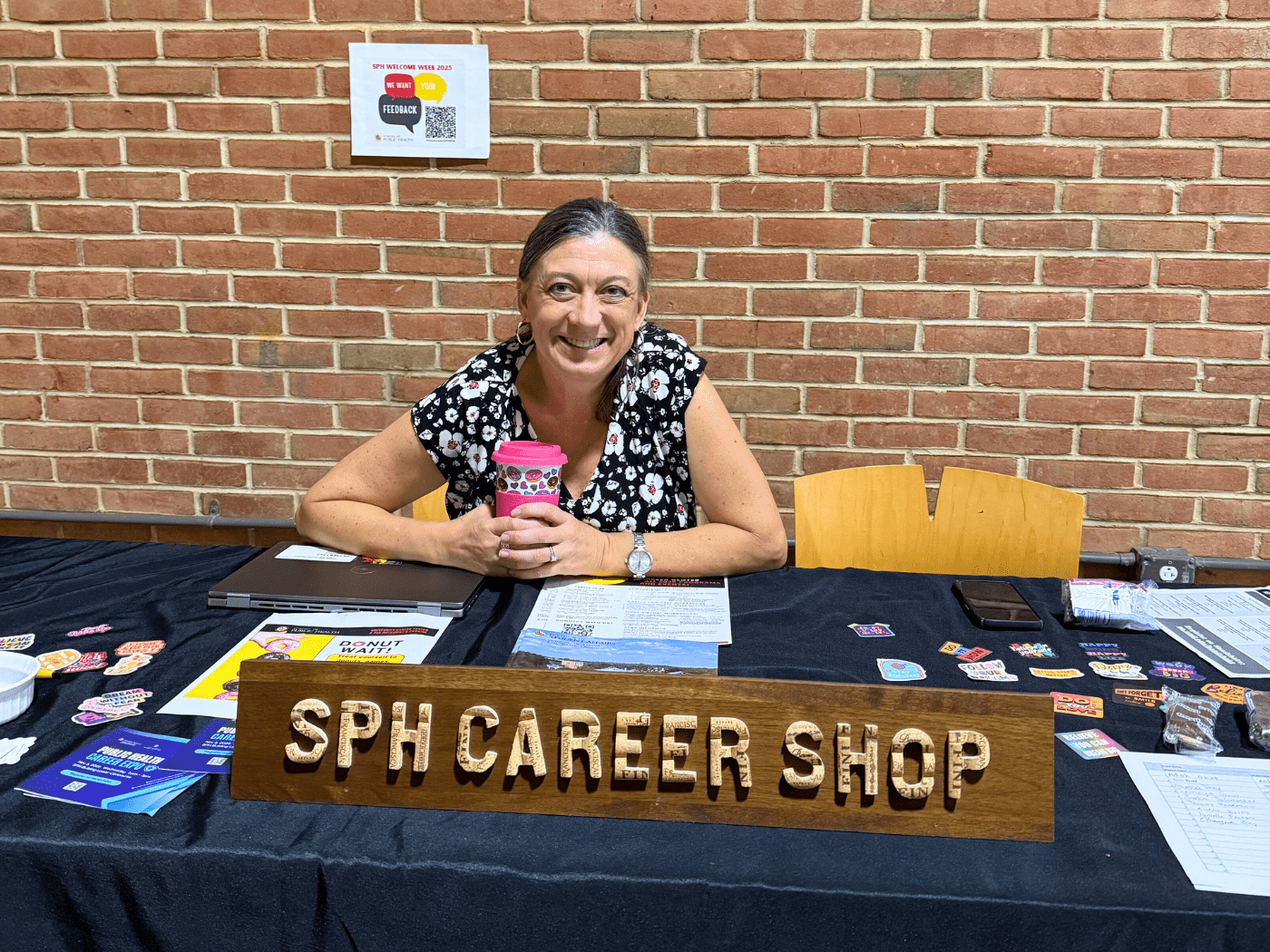 white woman sits at a table with sign for SPH Career Shop, wearing black and white floral shirt, smiling at camera