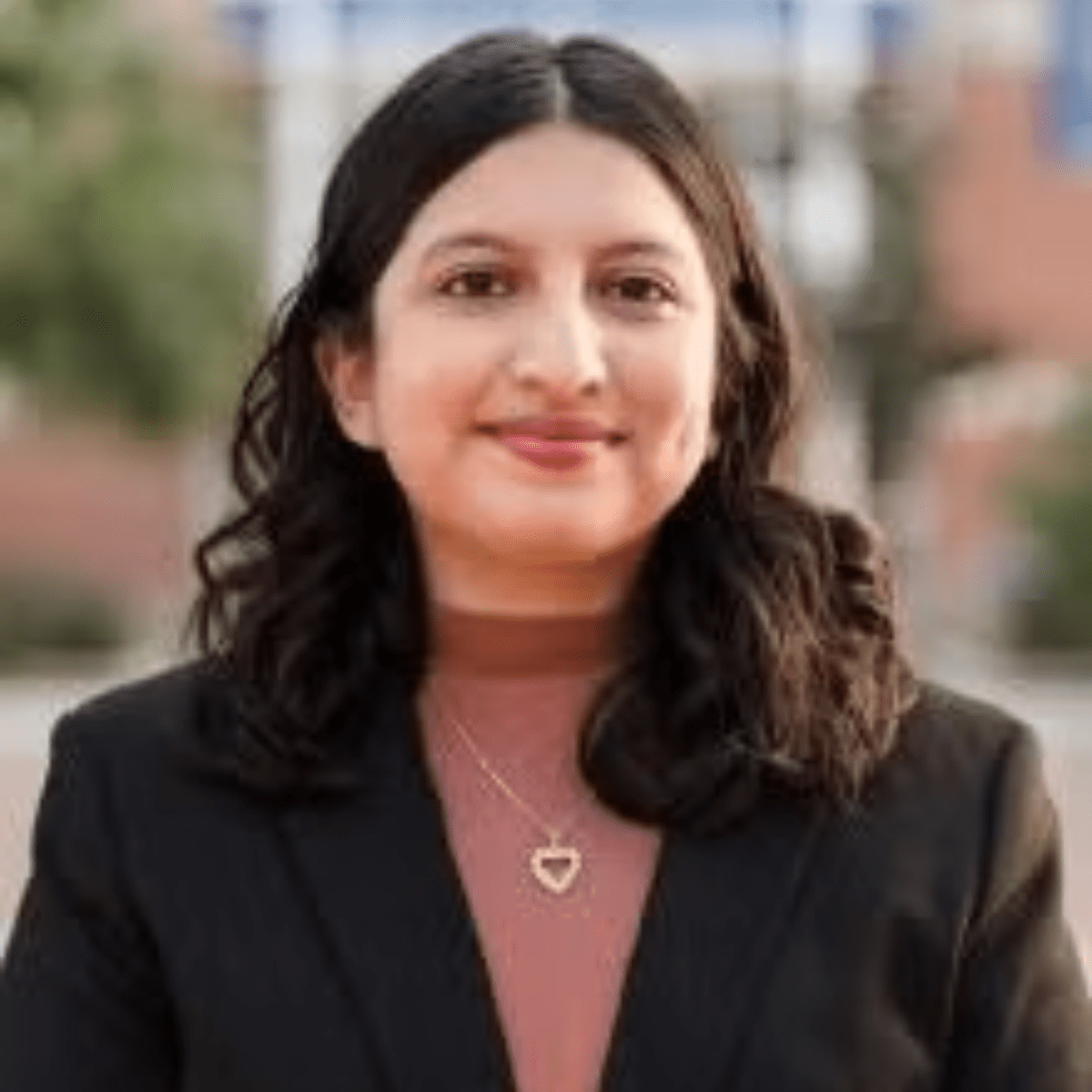 Headshot of a person with shoulder-length dark hair wearing a black blazer and a necklace, standing outdoors.