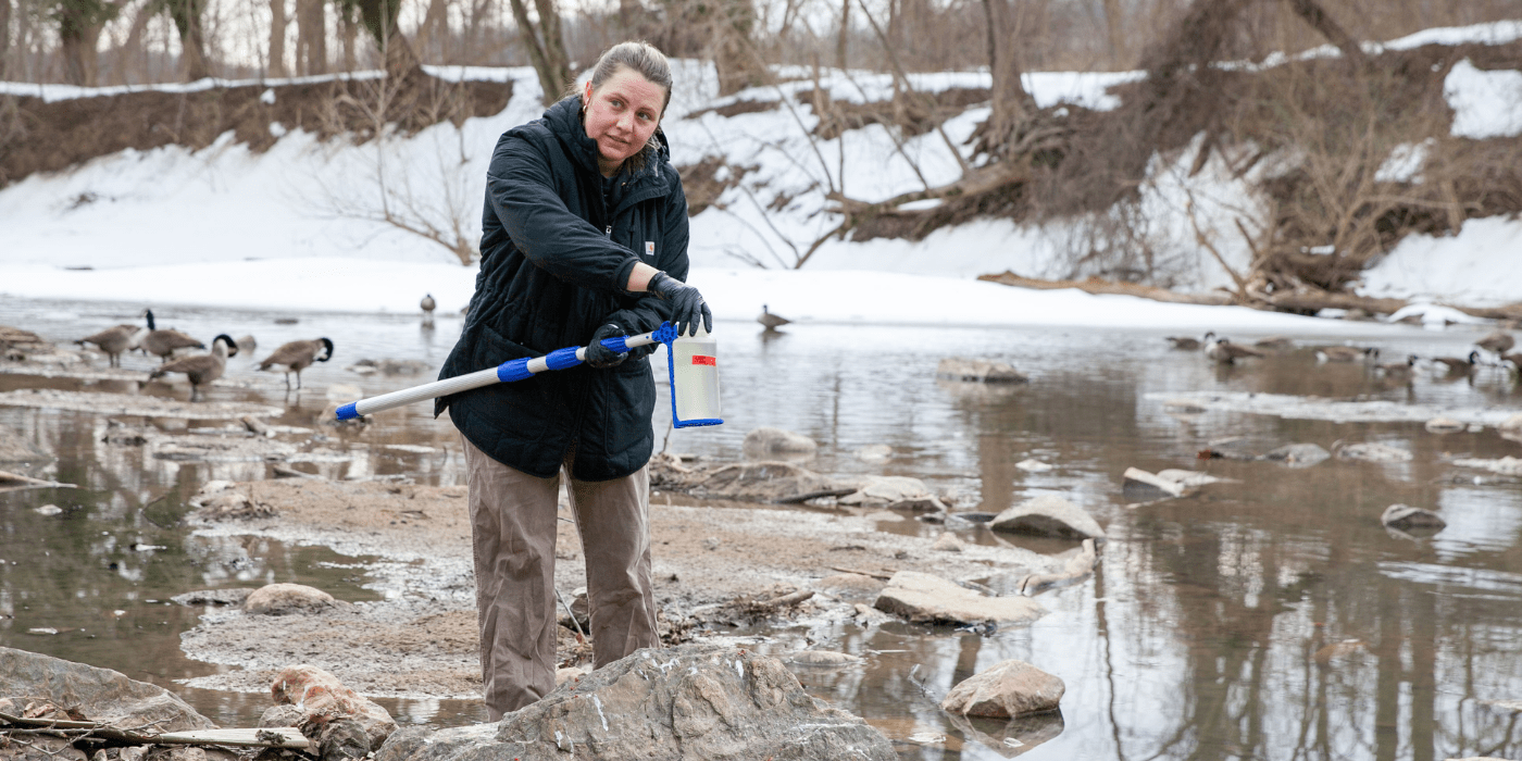 Person in a winter jacket uses a long-handled sampler to collect water from a creek with snow along the banks.