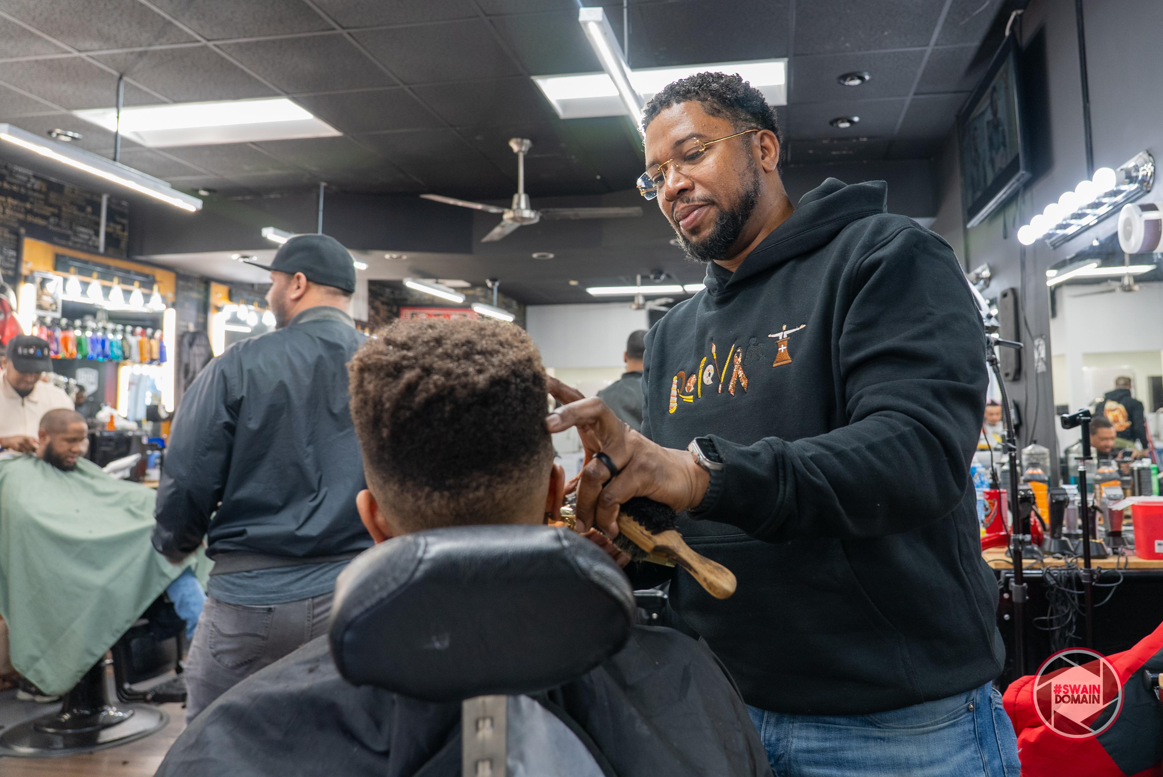 Black male barber cuts the hair of a Black man, sat in the chair with back of head to camera, barbershop in background with fluorescent lighting and other barbers and custoimers