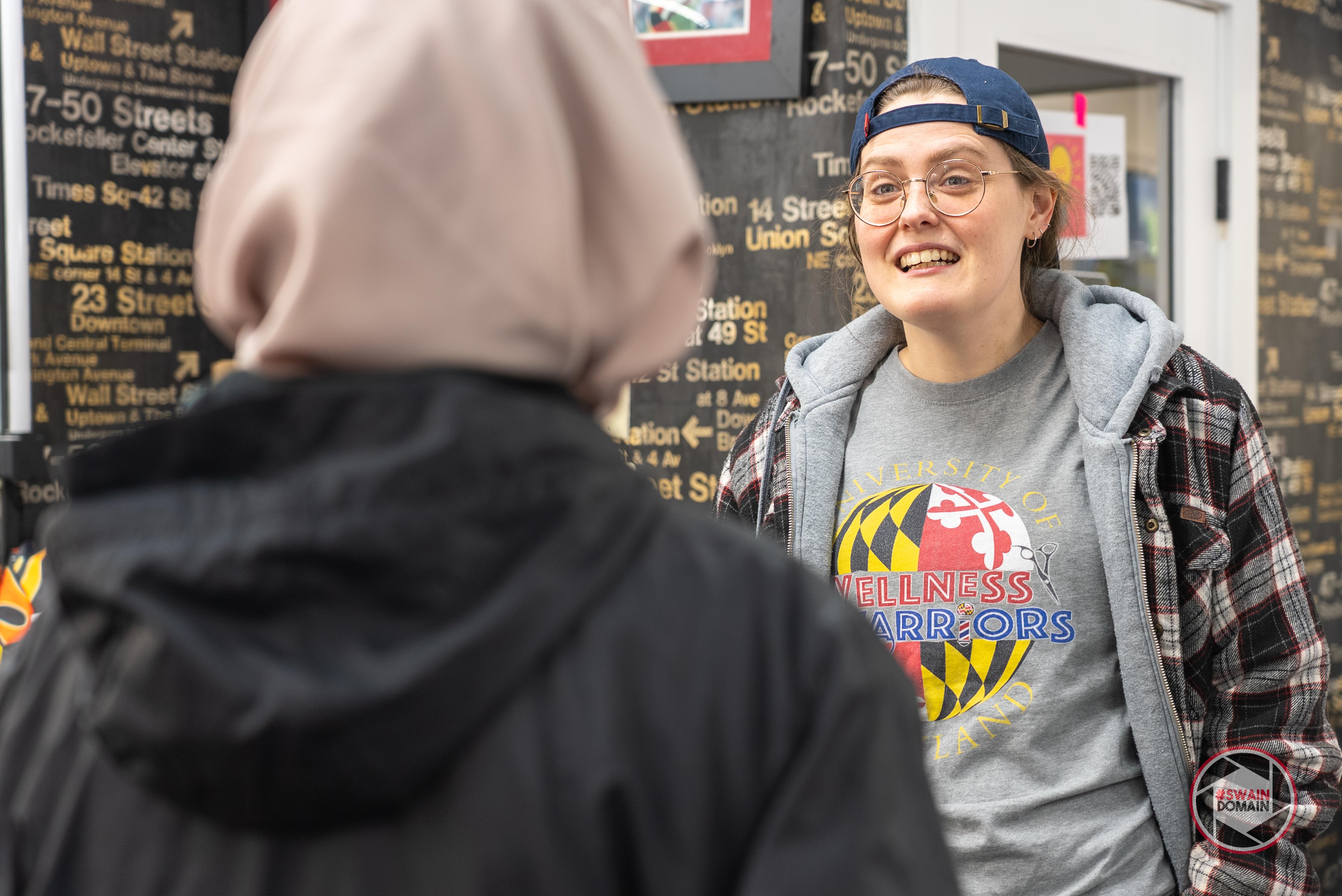 White person with backwards hat and a grey tshirt and plain shirt speaks to a student with a beige head covering and black hoodie, seen from behind, with barbershop wall in background
