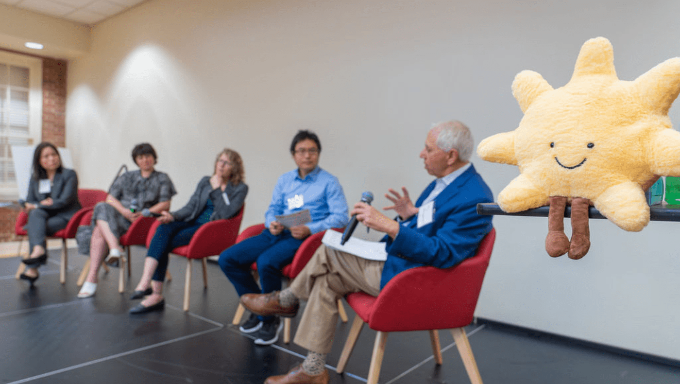 Five panelists sit in red chairs on stage, in foreground is a plushy of a yellow sun