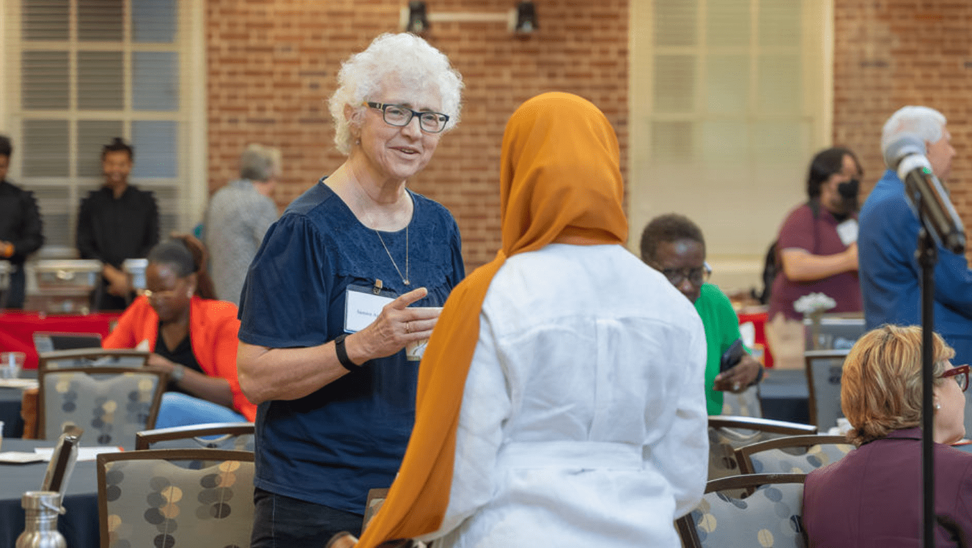 Older woman talks to another woman wearing aheadscarf, seen from behind in a conference room, with tables and people in background
