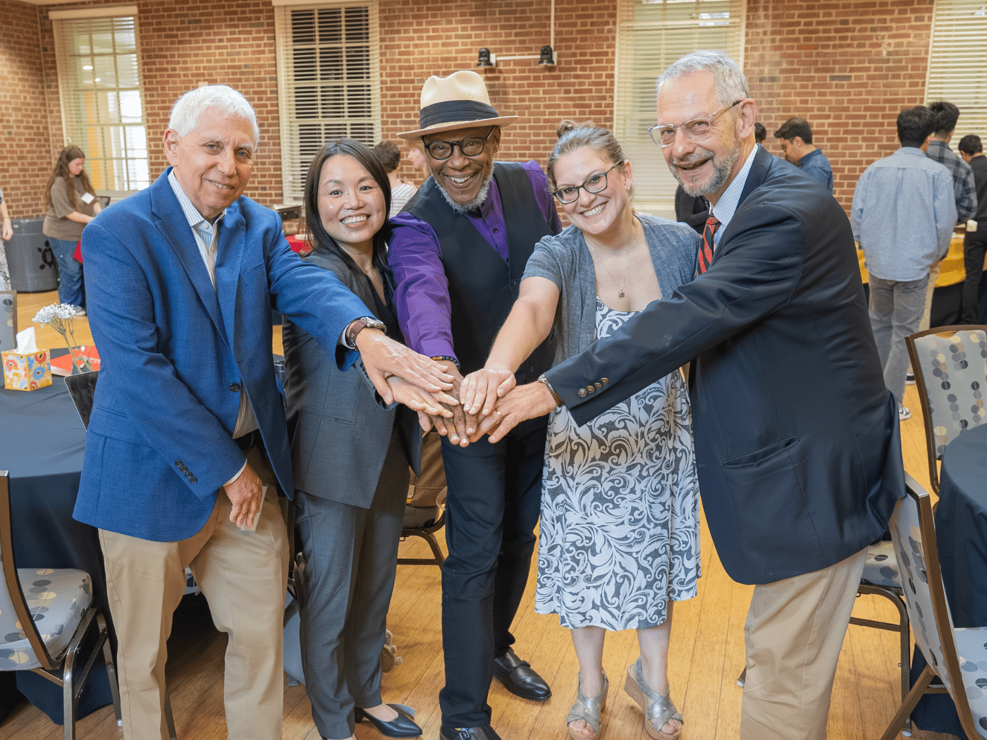 group of publihealth researchers pose with one arm reached out in front to touch hands, smiling, in a brick walled conference room