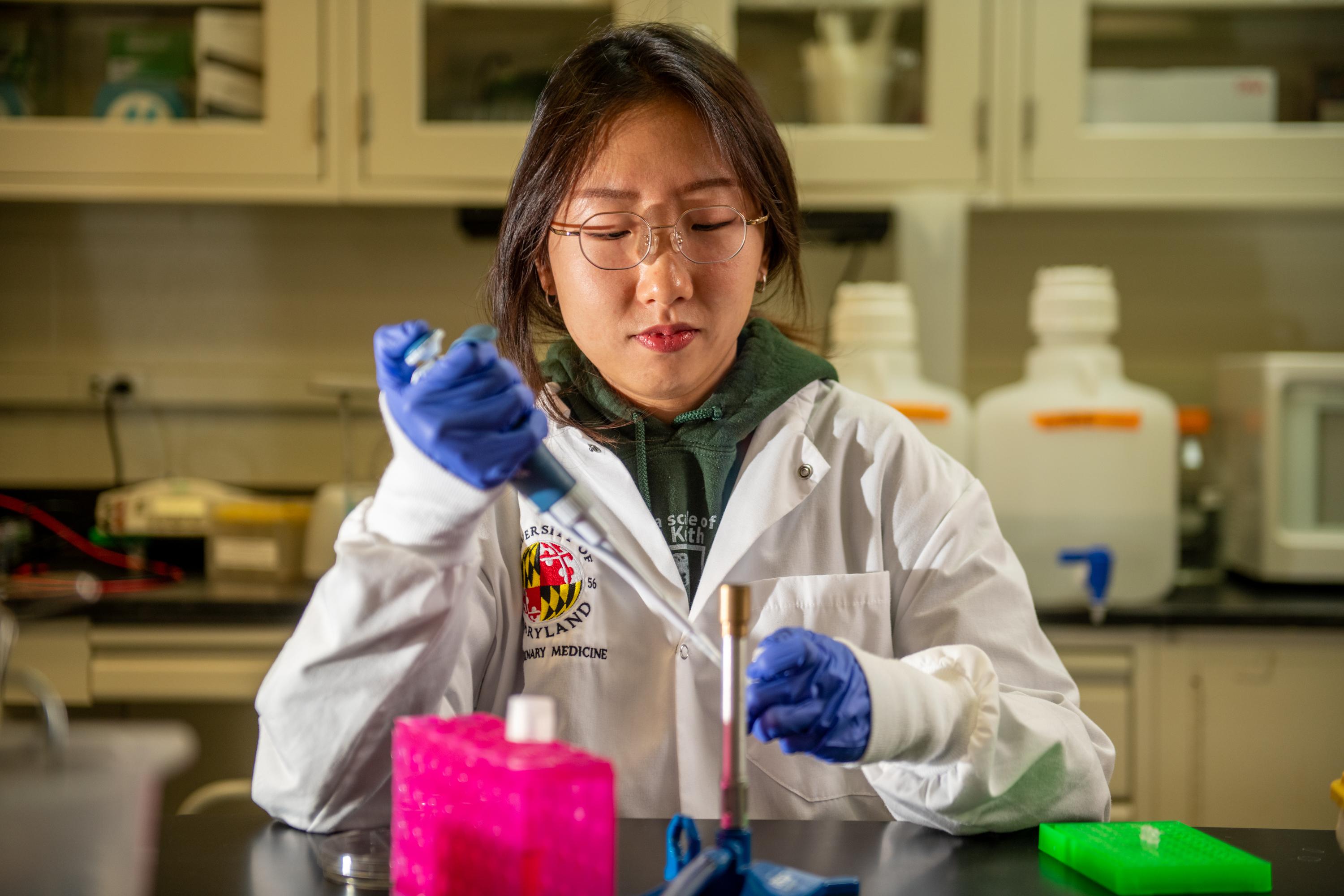 Asian student in white lab coat looks down at the syringe she is using to insert something into a small bottle, behind her shelves and materials of a biology lab