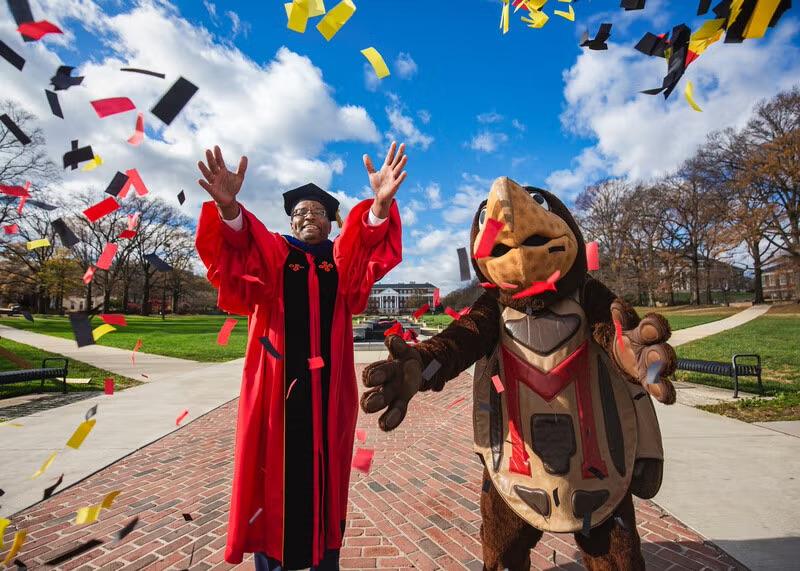 Darryll Pines, president of the University of Maryland, stands with Testudo on McKeldin Mall tossing red, black, and gold confetti, with campus buildings and a bright blue sky in the background.