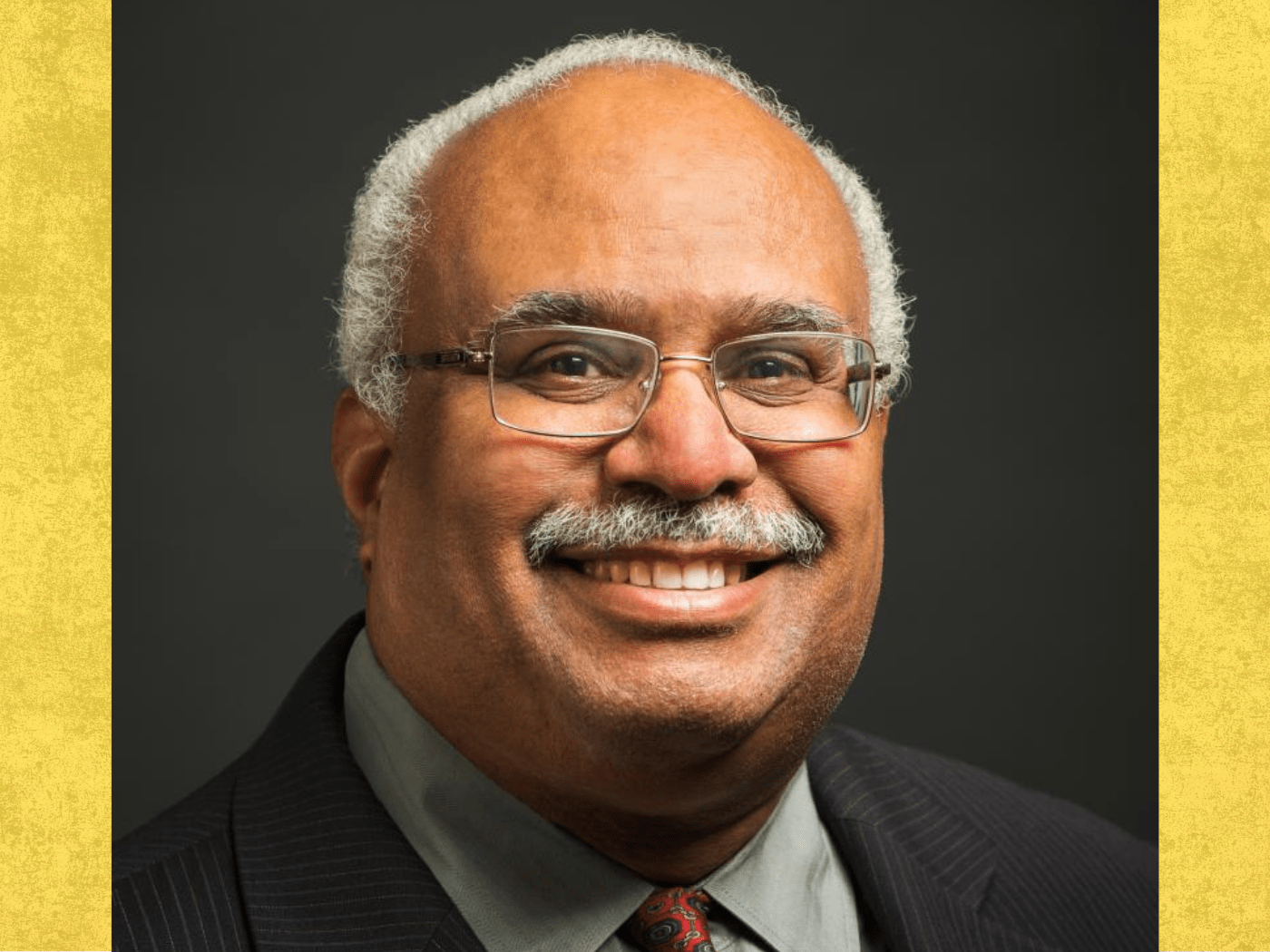 Black older man with greying moustache and rimless spectacle smiles to camera, wearing a dark suit jacket and reddish tie