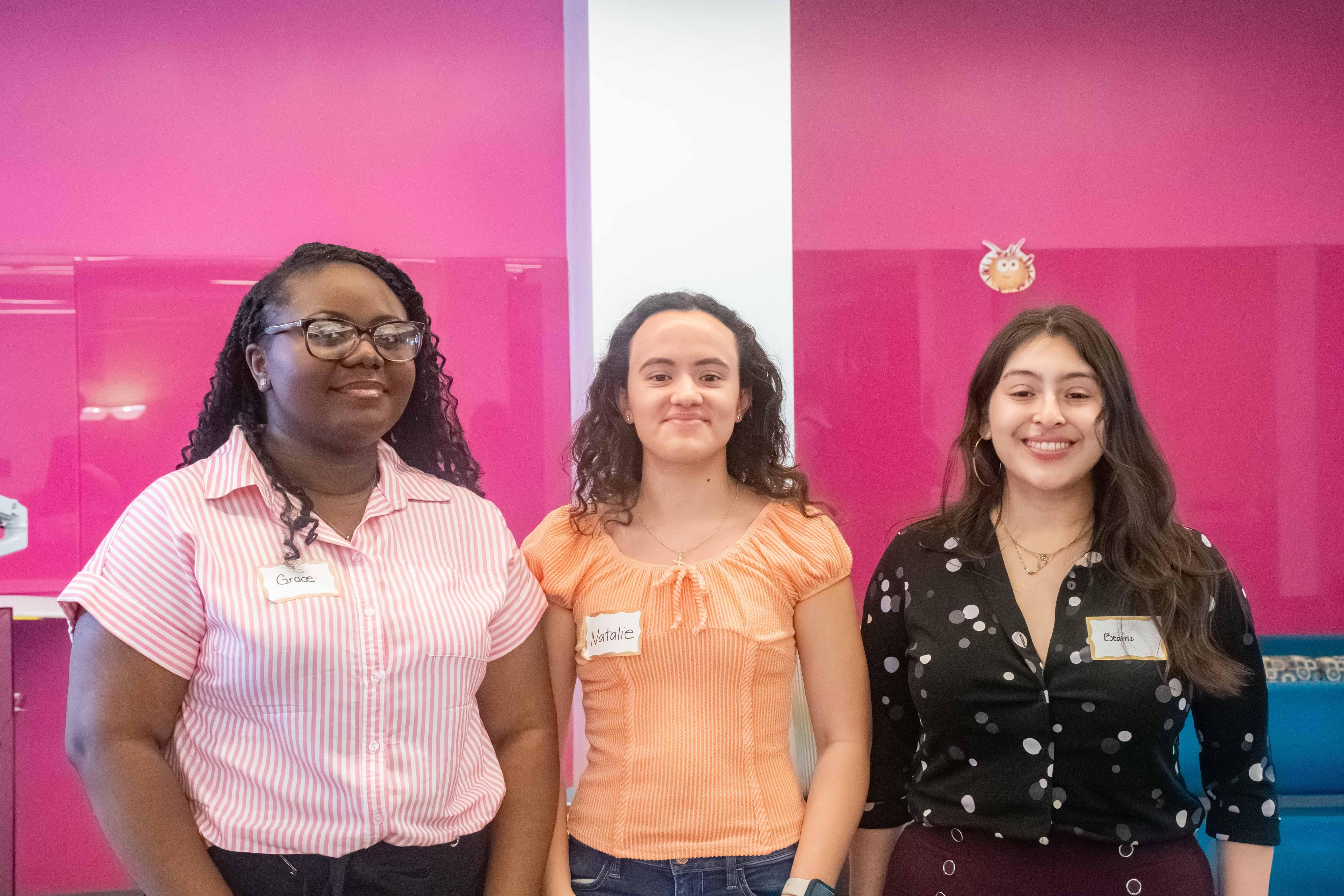 Three female students wearing pink shirt, orange top, black shirt respectively smile to camera in front of a pink background