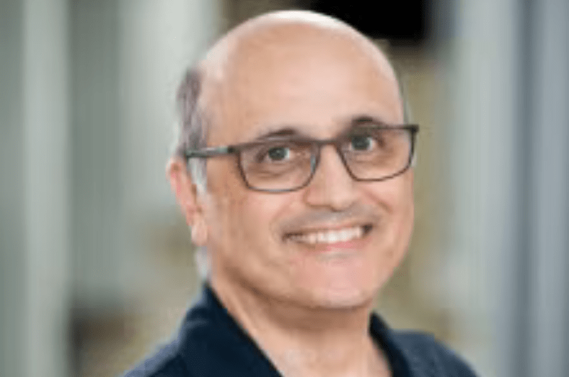 Headshot of a smiling bald man with glasses, wearing a dark collared shirt, against a blurred indoor background.