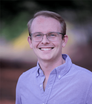 A medium close-up headshot photo of a light-skinned man wearing a purple button-up shirt and clear-rimmed glasses, with light-colored hair, smiling against a dark blurred background.
