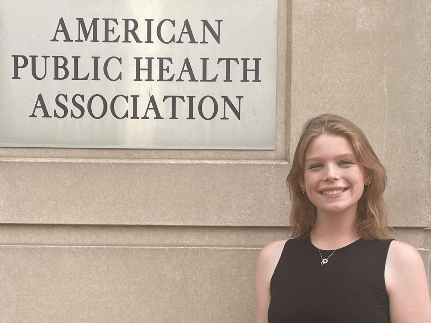 Young white woman in black tank top smiles to camera next to sign for the American Public Health Association
