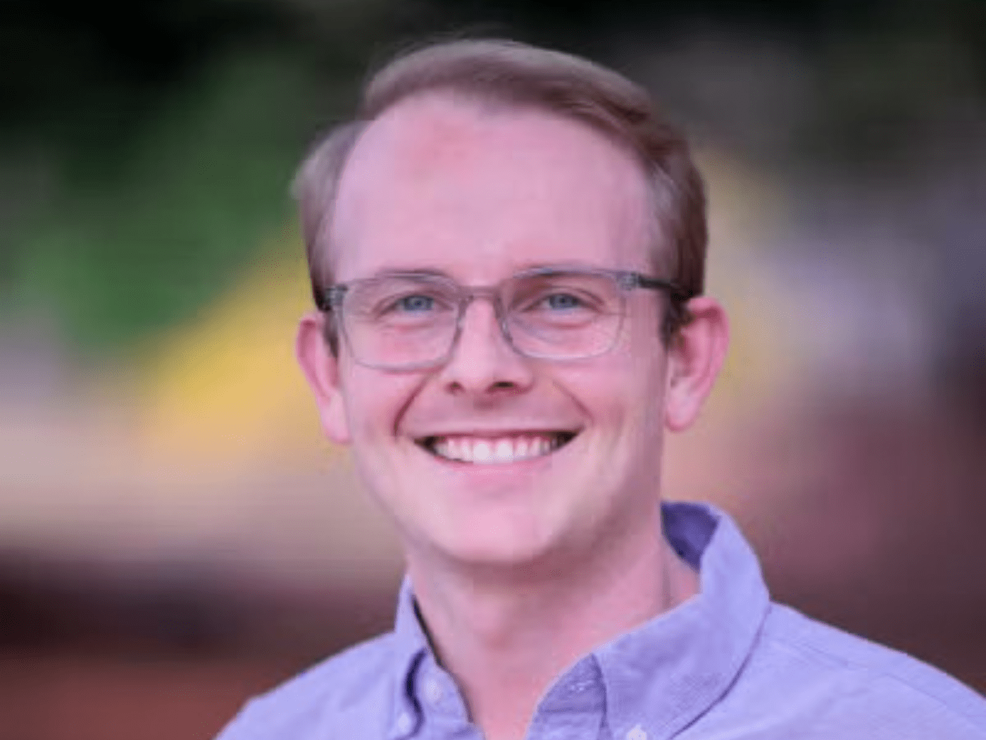 Man in purple button down smiles to camera wearing glasses