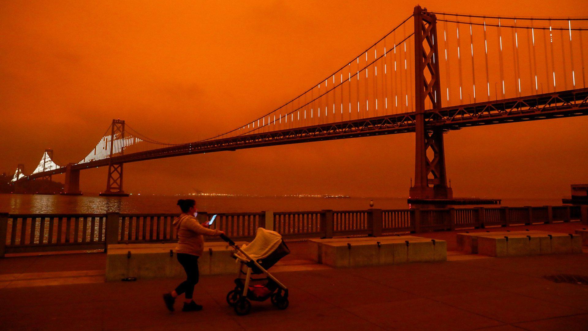 red smokey sky behind a big bridge, in foreground a person pushes a stroller while looking at their phone