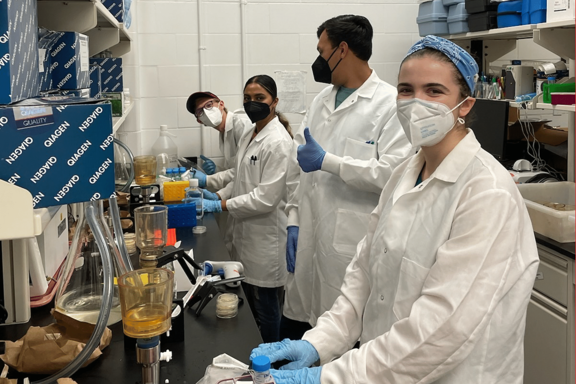 Four people all wearing white lab coats stand in a line at a lab table, wearing masks and holding the tools they use to test water samples