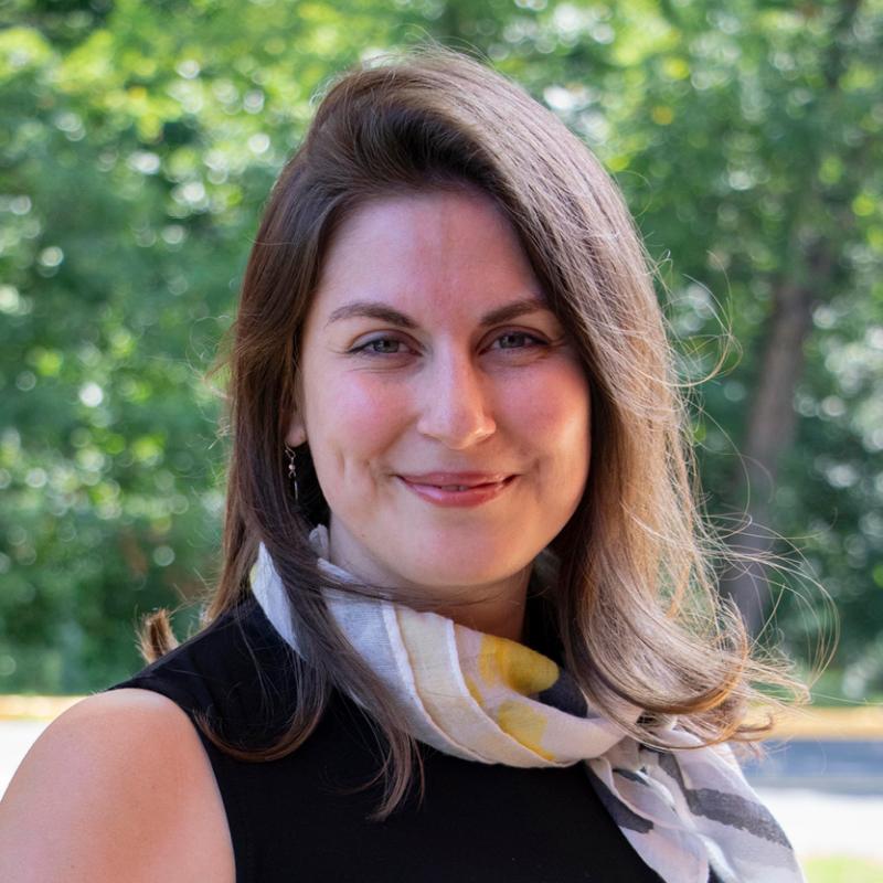 Headshot of a smiling person with long hair wearing a scarf, outdoors with greenery in the background.