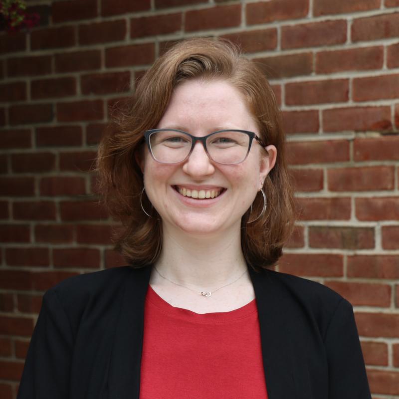 Smiling person wearing glasses stands in front of a brick wall.