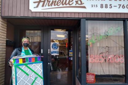 Masked woman stands outside Arnette’s Barber Shop holding a colorful decorated box.