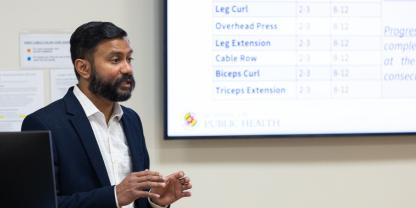 South Asian man with beard and dark hair in a dark suit with white shirt speaks, with hand gestures, in background the screen lists different kinds of resistance training types - eg bicep curls