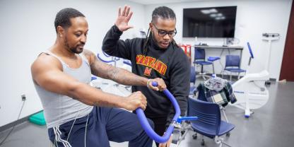 A black man in sweats rides a stationary bike, he has wires attached to him and another Black man stands beside him, wearing Maryland sweatshirt and glasses, he looks at the bike's monitor and counts of seconds with his raised hand